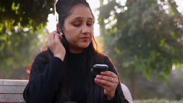 Closeup of an Indian woman wearing an earpod in her ear while sitting in a park and looking on camera. Calm outdoor lifestyle moment showing technology, focus, and modern daily life.