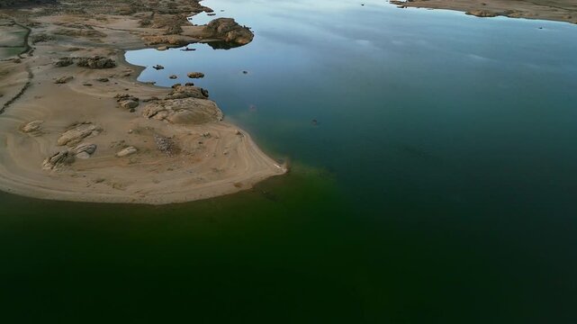 Aerial drone birds-eye view over Almendra Reservoir with clear lake water, visible sky reflections, and rocky sandy shoreline in Spain