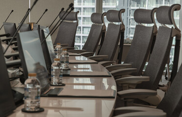 The modern conference room features a large, curved table set up with individual stations, each equipped with a personal screen, microphone, and water bottle. Sleek, high-back grey chairs are arranged