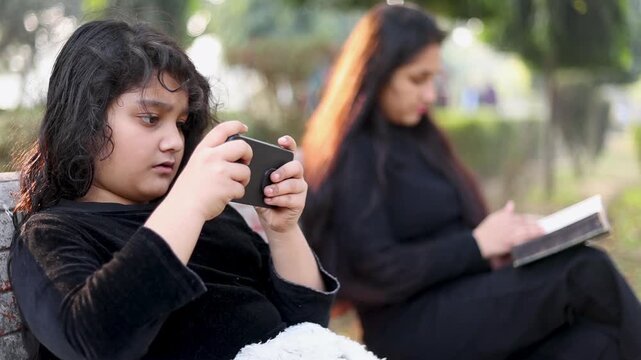 Closeup of an Indian child using a mobile phone while her mother reads a book on a park bench. Modern parenting moment showing screen habit, distance, and daily lifestyle contrast.