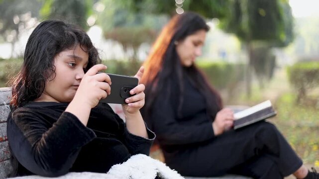 Closeup of an Indian child using a mobile phone while her mother reads a book on a park bench. Modern parenting moment showing screen habit, distance, and daily lifestyle contrast.