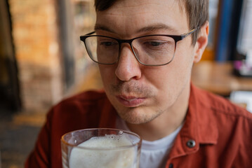Young man wearing glasses and a terracotta shirt is blowing gently on the foam of his beer in a pub, savoring the moment before taking a sip
