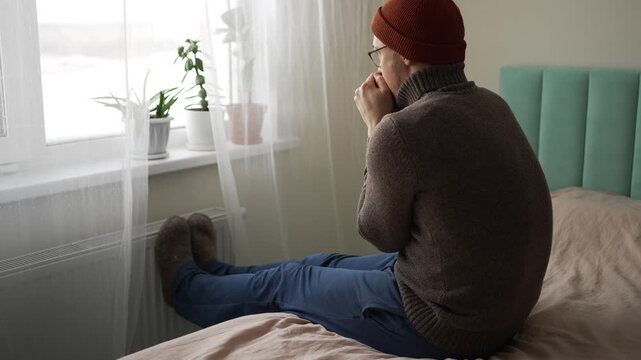 Unhappy shivering man wearing a warm sweater, hat, and socks sitting on a bed, warming his hands and feet on a central heating radiator due to the high cost of heating in winter