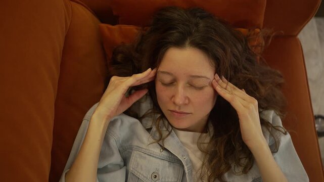 Overhead shot of a young woman dealing with a severe headache, resting on the sofa and gently rubbing her temples. An exhausted individual feeling unwell, enduring the discomfort of a migraine