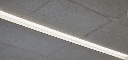 Fototapeta premium A bright linear LED light fixture mounted on a textured concrete ceiling with visible cracks and geometric patterns.