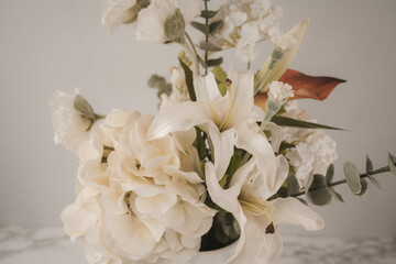 A beautiful bouquet of white flowers is arranged in a clear glass vase. The elegant display features lilies, hydrangeas, and lush greenery, placed on a smooth marble surface