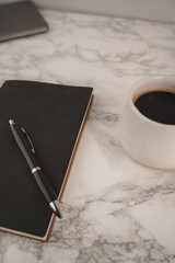 A black notebook and pen rest on a smooth marble table beside a white coffee cup filled with dark coffee. This scene captures a peaceful moment for creativity