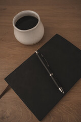 A black notebook and pen rest on a smooth marble table beside a white coffee cup filled with dark coffee. This scene captures a peaceful moment for creativity