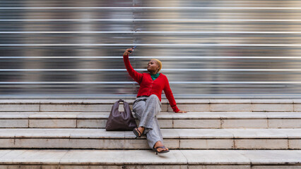 Young woman sitting on urban stairs taking selfie with extended reality glasses