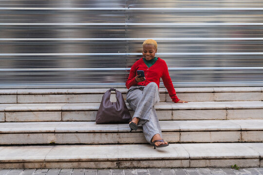 Young black woman sitting on stairs using a smartphone, enjoying social media or communication - Powered by Adobe