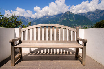 Wooden bench on a sunny terrace overlooking alpine mountains under blue sky and clouds, creating a serene outdoor scene with copy space.