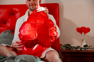 Man inflating heart-shaped balloons in a bedroom on a red bed, preparing a romantic setting for Valentine’s Day.	
