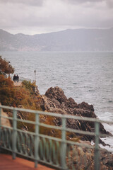 A couple strolls along a path by the rocky coastline. Waves crash against the rocks below. The sky is gray and overcast with mountains in the distance and trees nearby