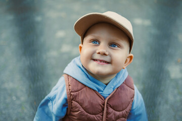 Little boy with bright blue eyes smiles joyfully in a cozy vest and cap during a sunny day at the park