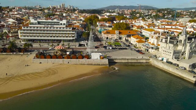 Drone reverse flight over sandy Cascais beach revealing Seixas Palace, red-roof town center and coastline on a bright sunny day.