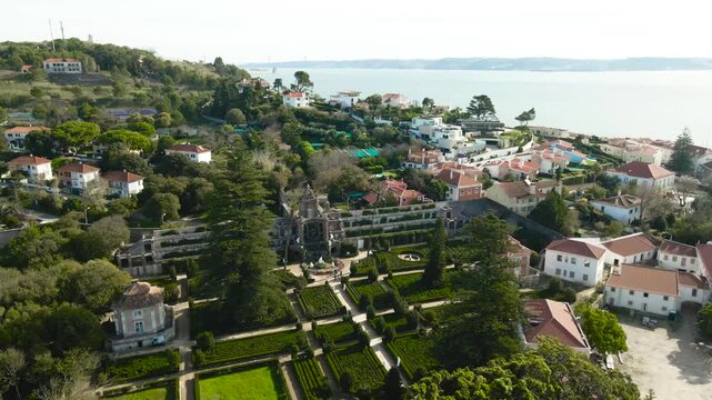 Drone orbit of Quinta Real de Caxias with symmetrical baroque gardens and Tagus River backdrop, surrounded by charming hillside houses on a sunny day.