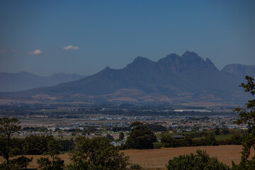 A wide-angle landscape view of the Stellenbosch region, featuring a sprawling valley with residential areas and agricultural fields set against a backdrop of dramatic, jagged mountain peaks.