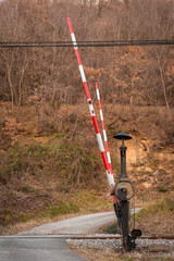 A traditional red-and-white railway crossing barrier stands raised beside a quiet rural road. Weathered metal mechanics and chains contrast with bare autumn trees and earthy hills in the background.