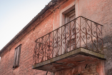 Weathered brick building with a rustic wrought-iron balcony and wooden door. Faded pink plaster, a shuttered window, and a clear sky create a nostalgic, Mediterranean atmosphere of quiet decay.