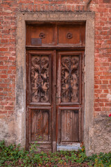 Weathered double wooden doors with ornate iron grilles featuring the initials "A" and "P." Set in a red brick wall with a stone frame, the entrance is surrounded by creeping greenery and history.