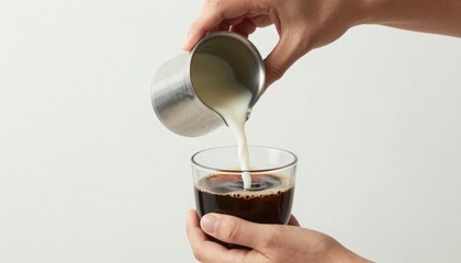 Close-up of human hands gracefully pouring fresh, creamy milk from a stainless steel pitcher into a clear glass filled with dark liquid, creating a delicious beverage mixture