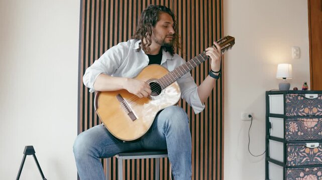 Young man with long hair sitting on a stool and emotionally singing a song while playing a classical acoustic guitar in a cozy room