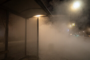 Dense steam from a burst central heating pipe covers a city bus stop at night, reducing visibility and creating an urban winter emergency scene.