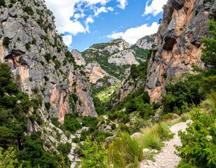 Deep canyon with weathered, colorful rock faces, lush vegetation, and a winding path under a partly cloudy blue sky