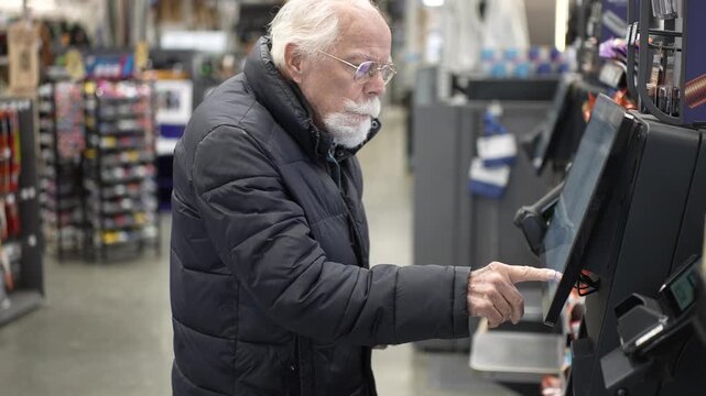An elderly man shops at a large hardware store and uses the self checkout to complete his purchase on a busy day.