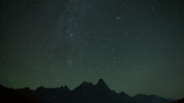 Night timelapse of the Milky Way and Andromeda Galaxy (M31) moving above the dark silhouette of Mount Ushba. Dramatic alpine night sky with clear celestial motion and mountain profile.