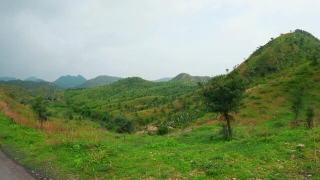 4K shot of green grass on Rayta hills and dark clouds in sky as seen from moving vehicle. Green forest in Sahyadri range at Udaipur, Rajasthan, India. Nature, environment and ecosystem concept.