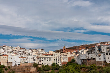 A panoramic view of Torrox Pueblo displays the dense white houses and the church tower against a blue sky. The traditional architecture of this Andalusian town rests on the hills of the Costa del Sol.