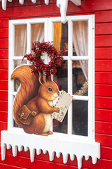 Christmas market decoration of a squirrel holding a letter in front of a red wooden house window with winter ornaments.
