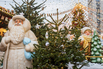 Moscow, Russia, 29 December 2025 Santa Claus and child winter festival statues among small decorated fir trees with festive string lights at a Christmas market.