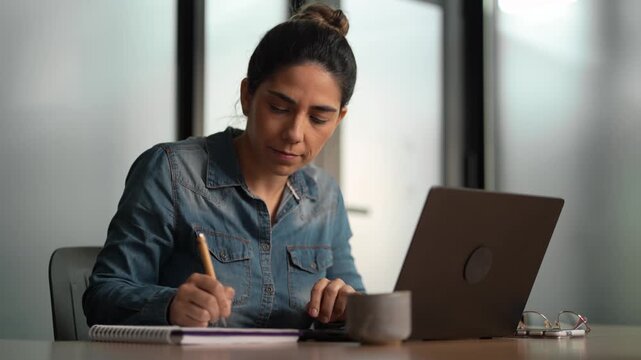 Title: Young professional woman in denim shirt taking notes in notebook while reviewing information on laptop at desk in modern office
