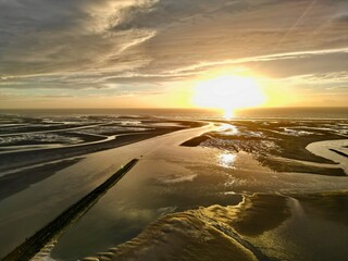 Drone aerial over Authie Bay tidal flats near Berck-sur-Mer, France, following a winding channel to the open sea at golden sunset under dramatic clouds and reflections