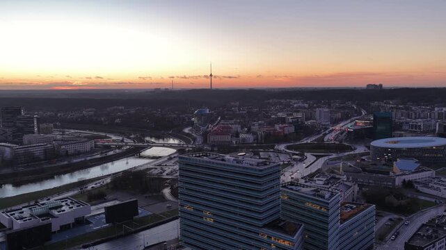 Panoramic aerial shot of Vilnius cityscape during sunset, showcasing modern skyscrapers, commercial buildings, and the iconic Vilnius TV Tower in the background.