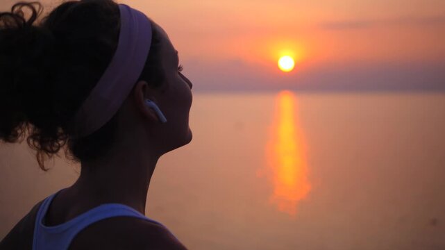 Young athletic woman in silhouette putting wireless earbuds in her ears while watching a beautiful sunset over the sea, getting ready for her evening run along the beach and enjoying the moment