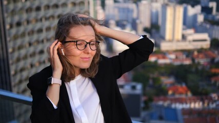 Relaxed young businesswoman with glasses and wireless earbuds enjoying music on a windy rooftop, with a beautiful city skyline and warm sunset light in the background