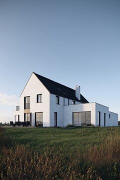 Modern house stands in a green field under a clear sky with simple design and large windows, offering a view of the surrounding landscape and bright sunlight during the day