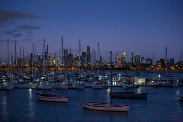 Sailboats are anchored in a bustling harbor as the Melbourne skyline glows during twilight hours.
