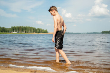 Young boy walks along the lake's edge, enjoying the warm sunshine. The clear sky and scenic nature create a peaceful, relaxed atmosphere at the water's edge on a summer day.