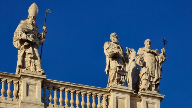Rome, Italy - 14 January 2025. Rooftop statues on San Giovanni in Laterano depict a bishop and two saints with a staff, book, and cross above a balustrade under clear skies.