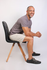Fototapeta premium Friendly mature man leaning on a chair smiling at camera, relaxed pose, gray t-shirt, studio portrait.