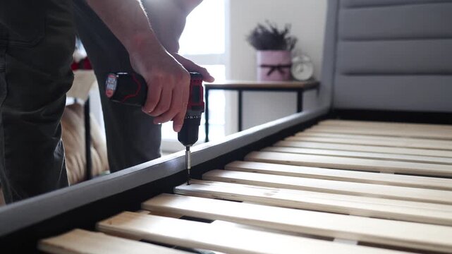 Self-assembly using a guide. House design. Close-up of a man's hands assembling a wooden bed using a screwdriver to insert screws into the bed frame.Collection of wooden furniture. Husband for an hour