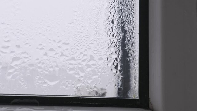 Damp window is covered in heavy condensation and water droplets. Macro shot captures the texture of moisture as the camera slowly tilts upwards.
