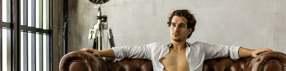 Banner. Young sexy handsome fit man in a brown suit looking at the camera sitting on a chair in the studio. Grey background, loft interior, studio. Causal stylish men's clothes, watch