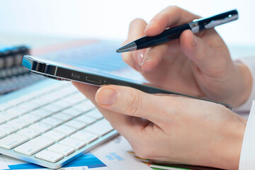 A woman works with a smartphone in the office, on a report. Close-up of her hands. 	