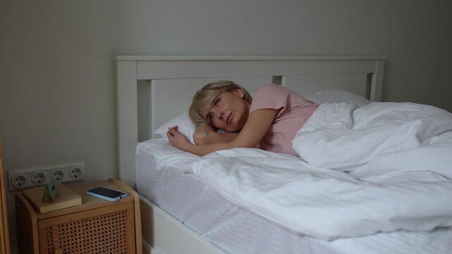Restless middle aged woman sleeping in bed, waking up after sleepless night, checking time on smartphone with tired unhappy expression, lazy to get up and falling back to sleep, insomnia and fatigue.