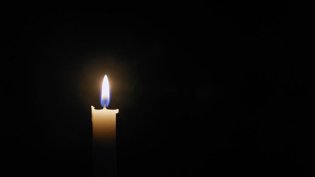 Single white candle burns with a flickering flame before being blown out, leaving complete darkness. The solitary light source creates a dramatic, high-contrast effect against the pitch-black